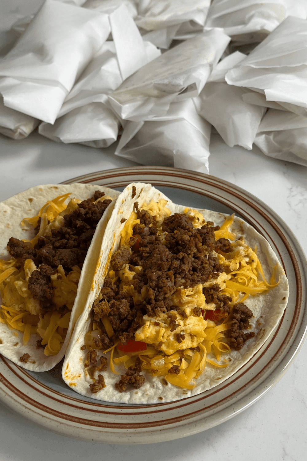 2 plated breakfast tacos with a bunch of wrapped breakfast sandwiches behind